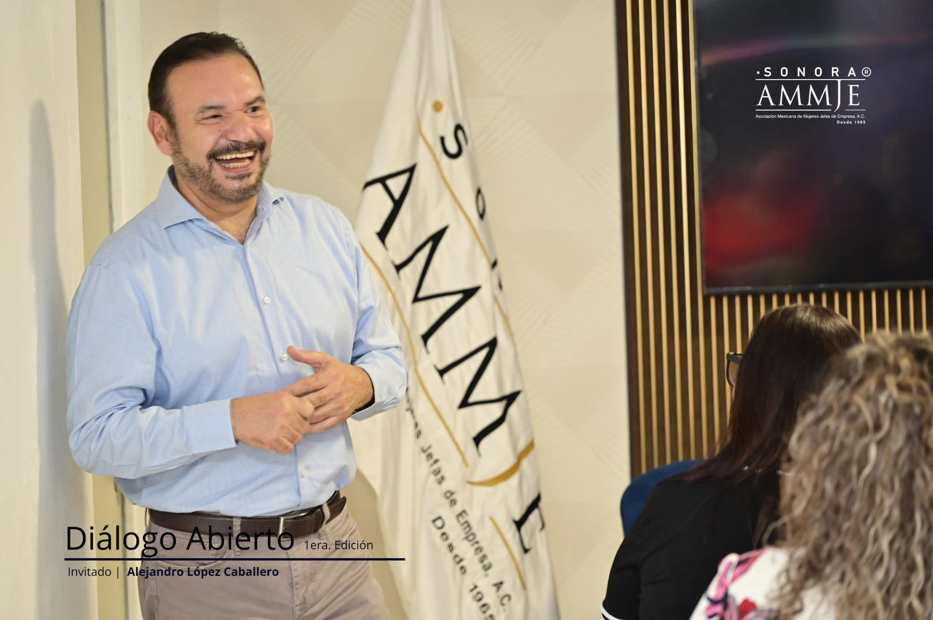 Alejandro López Caballero sonriendo junto al lábaro de AMMJE Sonora durante su participación en Diálogo Abierto